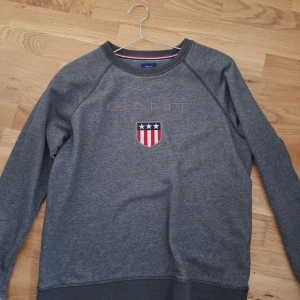 Grå sweatshirt från GANT - Säljer en stilren grå sweatshirt från GANT med ett broderat emblem på bröstet. Tröjan har långa ärmar och rund halsringning. Perfekt för en avslappnad stil. Den är helt ny, anledningen för att jag säljer den är att den är för liten och sitter bara i garderoben. Priset kan alltid diskuteras!