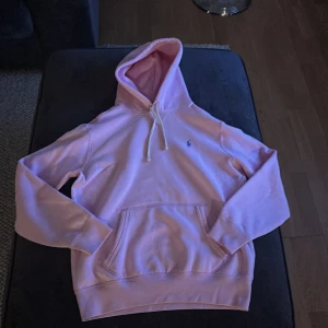 Rosa hoodie från Polo Ralph Lauren - Säljer en snygg rosa hoodie från Polo Ralph Lauren med en broderad logga på bröstet. Hoodien har en klassisk känguruficka och justerbar dragsko i huvan. Perfekt för en avslappnad stil!