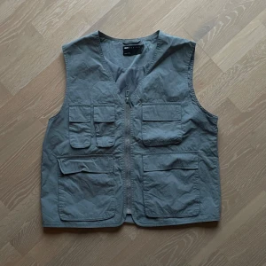 Gråblå Cargoväst från ASOS - Säljer en gråblå Cargoväst från ASOS. Den har en cropped/boxy fit och många snygga fickor. Västen har använts på danstävlingar vilket kan vara en rolig ”conversation starter”. 