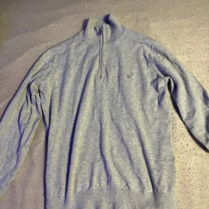 Grå tröja med dragkedja från GANT - Säljer en stilren grå tröja från GANT med halv dragkedja och broderad logotyp på bröstet. Perfekt för en avslappnad och snygg look. Tröjan har långa ärmar och är tillverkad i ett mjukt material. Storlek 158/164cm (13-14 år)