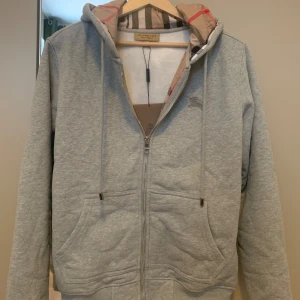 Grå hoodie från Burberry - Säljer en stilren grå hoodie från Burberry med dragkedja och klassiskt rutigt mönster i huvan. Tröjan har en broderad logga på bröstet och är perfekt för en avslappnad look. Passar utmärkt till både vardag och mer avslappnade tillfällen.