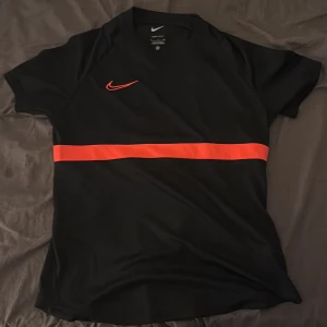 Svart Dri-FIT t-shirt från Nike - Säljer en svart Dri-FIT t-shirt från Nike med en röd rand över bröstet och ryggen. T-shirten har korta ärmar och är perfekt för träning. Nike-loggan är broderad i rött på framsidan.