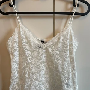 Vit spetslinne - Säljer ett vackert vitt spetslinne med tunna axelband. Perfekt för en somrig look eller att bära under en kavaj. Linnet har ett blommigt mönster och en v-ringad design.