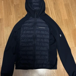 Mörkblå cardigan från Peak Performance - Snygg mörkblå cardigan från Peak Performance med quiltad framsida och huva. Jackan  har en dragkedja framtill och långa ärmar. Perfekt för kyligare dagar.