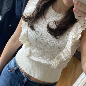 Vit ärmlös topp med volangdetaljer - Säljer en söt vit ärmlös topp med vackra volangdetaljer. Toppen har ett fint hålmönster och en ribbad nederkant som ger en snygg passform. Perfekt för varma dagar! I xs/s