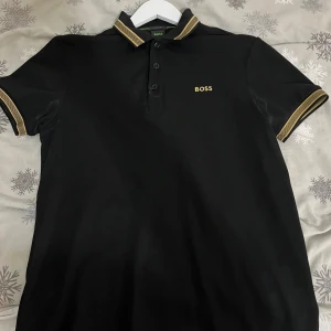 Svart pikétröja från Boss - Snygg svart pikétröja från Boss med guldfärgade detaljer på krage och ärmslut. Tröjan har en klassisk pikédesign med knappar framtill och korta ärmar. Perfekt för en stilren look. Pris kan diskuteras!!