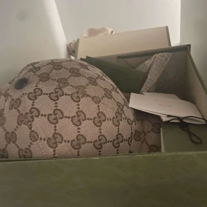 Gucci keps - Snygg beige keps från Gucci med det klassiska monogrammönstret i brunt. Perfekt för att ge en lyxig touch till din outfit. Kommer i originalförpackning.