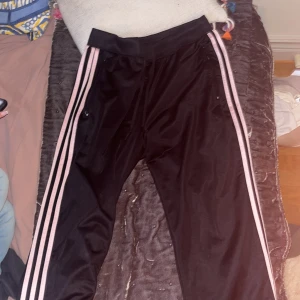 Svarta byxor från Adidas - Svarta träningsbyxor från Adidas med de klassiska tre ränderna i rosa längs sidorna. Byxorna har en elastisk midja och är tillverkade i ett syntetmaterial. Perfekta för träning eller en avslappnad dag.