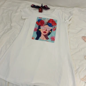 Vit t-shirtklänning med färgglatt tryck från Shein - Säljer en superfin vit t-shirtklänning från Shein. Den har ett stort, färgglatt tryck på framsidan med en cool illustration av en kvinna med blommor i håret och solglasögon. Perfekt för en avslappnad sommarlook! Klänningen är kortärmad och gjord i ett mjukt material som känns skönt mot huden.