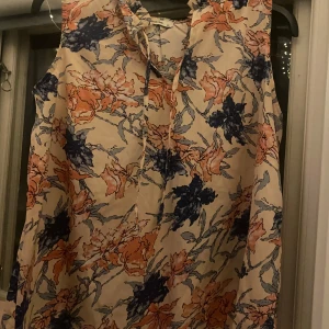 Blommig ärmlös topp från ChaCha - Säljer en superfin ärmlös topp från ChaCha i storlek XL. Den har ett härligt blommigt mönster i blått och orange på en beige bakgrund. Perfekt för sommardagar eller en avslappnad festkväll. Toppen har en knytning vid halsen som ger en söt detalj.