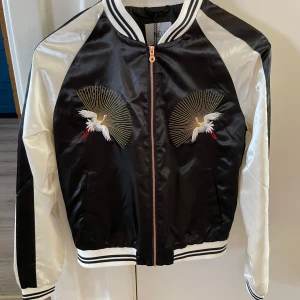 Svart och vit bomberjacka från NEU - Säljer en helt ny svart och vit bomberjacka från NEU. Jackan har ett vackert broderat mönster med fåglar på framsidan och är tillverkad i ett glansigt syntetmaterial.