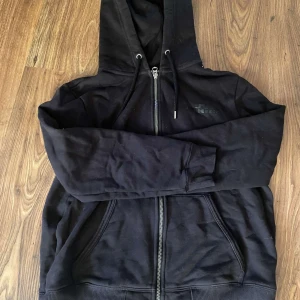Svart hoodie från J.Lindeberg - Säljer en stilren svart hoodie från J.Lindeberg. Den har en dragkedja framtill och justerbar huva med snören. Perfekt för en avslappnad look. Den är i dam storlek M så den är lite mindre än vanlig. Kontakta mig om du har några frågor.