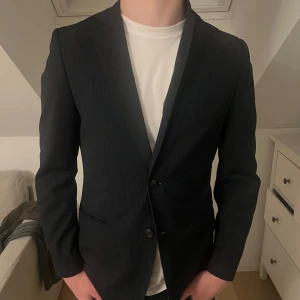 Svart kavaj - Snygg svart kavaj med smal passform och diskret randigt mönster. Perfekt för både formella och semi-formella tillfällen. Kavajen har två knappar framtill och en klassisk krage.