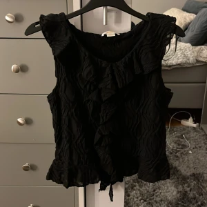 Svart topp från Gina Tricot - Säljer en snygg svart topp från Gina Tricot med volangdetaljer framtill. Priset kan diskuteras