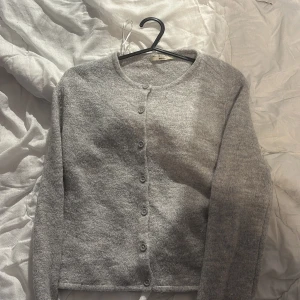 Grå kofta från Gina Tricot - Mysig grå kofta från Gina Tricot i premiumkvalitet. Den har långa ärmar och knappar framtill, perfekt för kyliga dagar. Enkel och stilren design som passar till det mesta.