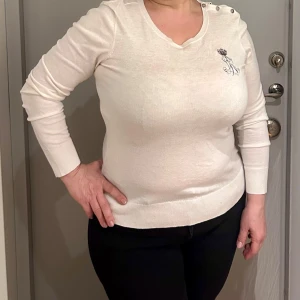 Vit långärmad tröja från Lauren - Elegant vit långärmad tröja från Lauren med broderad logotyp och krona på bröstet. Tröjan har dekorativa knappar på axeln som ger en stilren touch. Perfekt för en sofistikerad look.