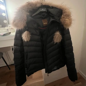 Svart dunjacka med pälsdetaljer från Bark of Sweden - Säljer en snygg svart dunjacka från Bark of Sweden med pälsdetaljer på huvan. Jackan har en dragkedja framtill och är perfekt för kyliga dagar🖤 Orginal pris är 2000kr