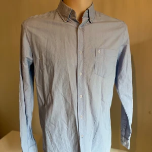Ljusblå skjorta med knappkrage - Snygg ljusblå skjorta med knappkrage och bröstficka. Skjortan har långa ärmar och knappar framtill. Perfekt för en stilren look.