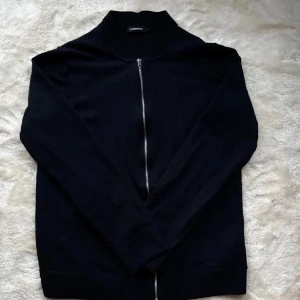 Svart tröja med dragkedja från J.Lindeberg - Snygg svart tröja från J.Lindeberg med dragkedja framtill. Perfekt för en stilren look. Tröjan har långa ärmar och en enkel design som passar till det mesta. Väldigt liten i storleken så den skulle passa S/M