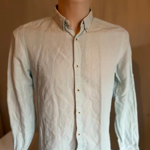 Ljusblå Linnéskjorta med knappar - Snygg ljusblå skjorta från Zara med klassisk krage och knappar hela vägen ner. Perfekt för en stilren look. Skjortan har långa ärmar och är tillverkad i ett lätt material som ger en skön känsla.