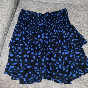 Blåmönstrad kjol - Snygg kortkjol med blått bladmönster på svart bakgrund. Kjolen har en elastisk midja för bekväm passform och volanger som ger en lekfull look. Perfekt för att liva upp garderoben med lite färg och mönster. Väldigt söt och romantisk.