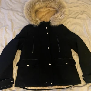 Svart parkajacka med pälsfodrad huva - Snygg svart parkajacka med en mysig pälsfodrad huva och quiltat foder. Jackan har knappar och dragkedja framtill samt praktiska fickor med lock. Perfekt för kyliga dagar.