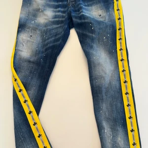Jeans med gula detaljer från Dsquared2 - Jeans från Dsquared2 med en unik design. De har en blå tvätt med slitningar och gula sidoremsor med svarta detaljer. Perfekta för dig som vill sticka ut med stil. Tillverkade i Italien. Storlek 46, nypris 7000:-, använda 2 ggr