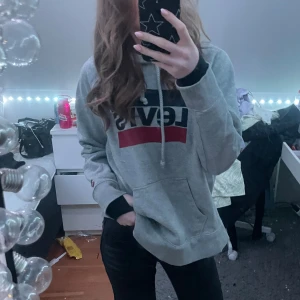 Grå hoodie från Levi's - Säljer en grå hoodie från Levi's med klassisk logga i svart och rött på bröstet. Tröjan har en stor ficka framtill och justerbar huva med snören. Perfekt för en avslappnad stil. 🖤