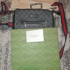 Säljer en stilren svart axelväska från Gucci med det klassiska GG-mönstret. Väskan har en justerbar axelrem i rött och blått, samt en praktisk dragkedja framtill. Perfekt för att bära dina viktigaste saker med stil. Vid frågor är det bara att skriva!