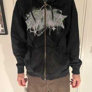 Svart hoodie från Weekday - Säljer en svart hoodie från Weekday med ett coolt grafiskt tryck i lila och grönt på framsidan. Hoodien har dragkedja och långa ärmar, perfekt för en avslappnad stil. Den har en klassisk passform och är gjord i mjukt material för extra komfort.