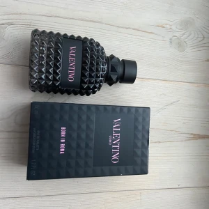 Valentino Uomo Born in Roma - Säljer en elegant parfymflaska från Valentino, modell Uomo Born in Roma. Flaskan har en unik design med ett mönster av svarta nitar och en stilren svart kork. Förpackningen är svart med rosa text. Perfekt för den som vill ha en sofistikerad doft.