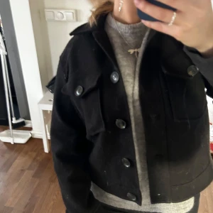Svart jacka med knappar - Snygg svart jacka med knappar framtill och två bröstfickor. Jackan har en klassisk krage och långa ärmar, perfekt för en stilren look. Passar bra till både vardag och mer uppklädda tillfällen.