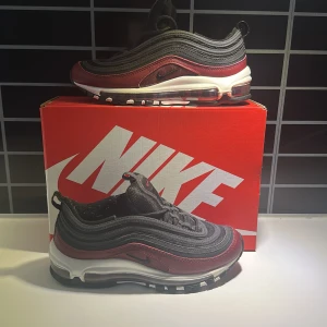 Nike Air Max 97 i svart och röd - Snygga Nike Air Max 97 sneakers i en cool kombination av svart och röd. Skorna har en vågig design med synlig Air-enhet i sulan för extra komfort. Perfekta för en sportig look med stil.