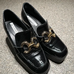 Svarta loafers från Blankens ”The Olivia”  - Snygga svarta loafers från Blankens med en elegant gulddetalj framtill. Skorna har en robust klack och en glansig finish som ger en lyxig känsla. Perfekta för att lyfta vilken outfit som helst.