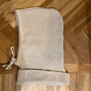 Beige stickad balaklava - Snygg beige stickad balaklava med ribbad design och knytband på sidan. Perfekt för kyliga dagar och ger en stilren look. Kragen går att vika upp och ned. 