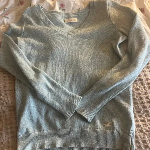 Ljusblå stickad tröja från Hollister - Mysig ljusblå tröja från Hollister med v-ringning och långa ärmar. Perfekt för kyligare dagar. Den har en diskret logga vid nederkanten och ribbade detaljer vid ärmslut och nederkant.