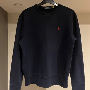 Mörkblå tröja från Polo Ralph Lauren - Snygg mörkblå tröja från Polo Ralph Lauren med det klassiska röda broderade logotypen på bröstet. Tröjan har långa ärmar och en rund halsringning. Perfekt för en stilren och avslappnad look. Tröjan är i storlek S och i väldigt bra skick. Bara att höra av sig om du har några eventuella frågor!☺️
