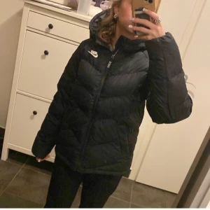 Svart dunjacka från Nike - Säljer en svart dunjacka från Nike med dragkedja och huva. Perfekt för kyliga dagar med sin pufferdesign som håller dig varm. Änvänt en gång så det är jätte bra skick 