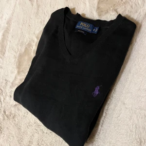 Svart tröja från Ralph Lauren - Snygg svart tröja från Ralph Lauren i pirma bomull. Tröjan har en v-ringad design och ett lila broderat logotyp på bröstet. Perfekt för en stilren look.