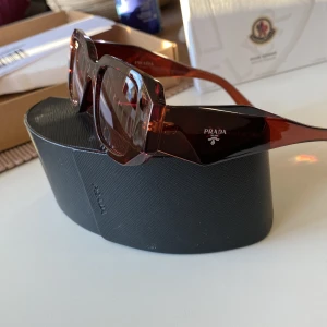 Bruna solglasögon från Prada - Snygga bruna solglasögon från Prada med rektangulära bågar. De har en elegant design med mörka linser och tydlig logotyp på sidan. Perfekta för att ge en lyxig touch till din stil.