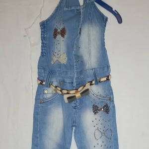 Jeansbyxdress med paljettdetaljer från White Cat - Snygg jeansbyxdress för barn från White Cat med paljettdetaljer i form av rosetter. Byxdressen har en smockad överdel och ett dekorativt bälte med kedjedetaljer. Perfekt för en stilfull look! Tyvärr vet jag inte vad size 1 betyder.