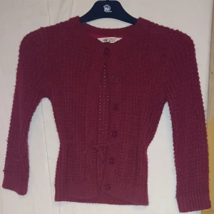 Vinröd barnkofta från H&M - Säljer en vinröd kofta från H&M med knappar framtill. Koftan har en stickad struktur och långa ärmar, perfekt för kyligare dagar. Passar barn i storlek 110/116.