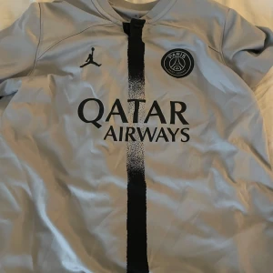 Grå PSG fotbollströja med Neymar Jr - Snygg grå PSG fotbollströja från Jordan med Neymar Jr och nummer 10 på ryggen. Tröjan har en svart vertikal rand på framsidan och Qatar Airways som sponsorlogga. Perfekt för fotbollsfans!