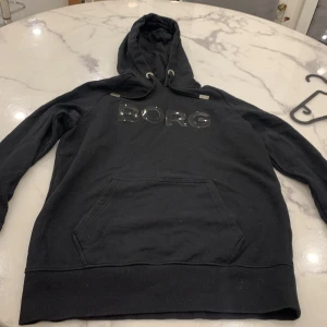 Svart hoodie från Björn Borg - Säljer en svart hoodie från Björn Borg med stor logga på bröstet. Tröjan har en klassisk känguruficka och justerbar huva med snören. 
