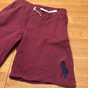 Vinröda shorts från Polo Ralph Lauren - Snygga vinröda shorts från Polo Ralph Lauren med en stor broderad logga i mörkblått. De har en bekväm passform med snörning i midjan. Perfekta för en avslappnad stil.