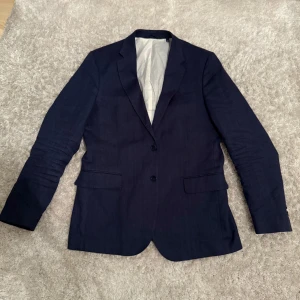 Mörkblå kavaj från GANT - Snygg mörkblå kavaj från GANT i slim passform. Kavajen har två knappar framtill och en stilren design. Perfekt för en elegant look. Framkommer ej storlek i kavajen men den passar mig som har M perfekt (har även L i vissa kläder).