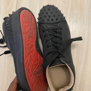 Christan Louboutin  - Snygga svarta sneakers med nitar på ovansidan och en röd sula. Skorna har snörning och en unik design som sticker ut. Köpt för 14000