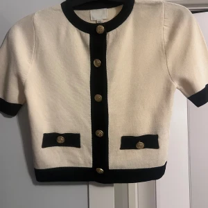 Vit och svart kofta med guldknappar - Kortärmad svart och vit/beige tröja med knappar den är aldrig använd och helt ny säljer den eftersom jag inte sett att den hade svarta fläckar på ryggen 