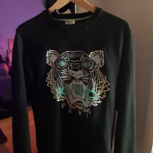 Svart tröja med tigertryck från Kenzo - Snygg svart tröja från Kenzo med ett iögonfallande tigertryck i silver och grönt på framsidan. Tröjan har långa ärmar och en rund halsringning. Perfekt för att ge din outfit en cool touch.