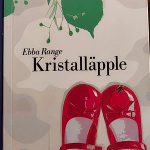 Kristalläpple - Boken 'Kristalläpple' av Ebba Range har ett omslag med en illustration av röda skor och gröna blad. Titeln är tryckt i svart mot en ljus bakgrund.
